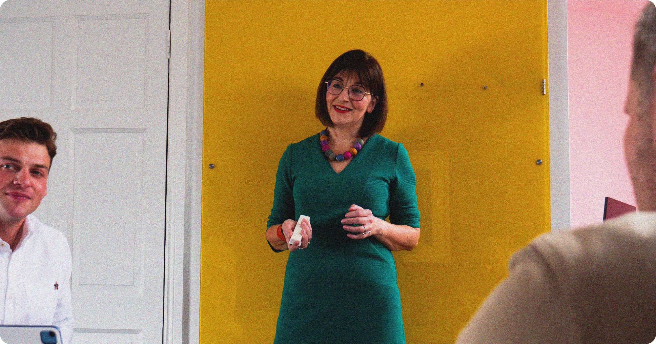 A woman stands in front of a yellow background, coaching a team on leadership and company culture.