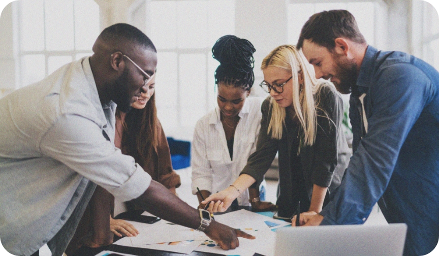 A group of diverse people lean over a desk, pointing at a document as they discuss organisational culture.