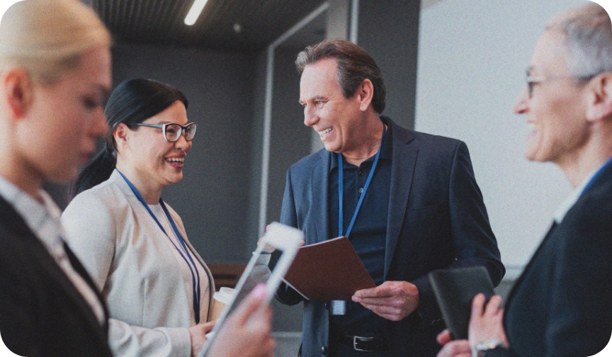 One man and three women stand around chatting. They are wearing lanyards and are dressed in business attire, holding clipboards and tablets, as they discuss workplace culture.