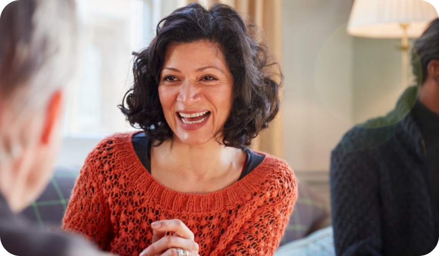 Woman with dark hair and burnt umber jumper smiles at two men who are visible partially from behind as they discuss defining company culture.